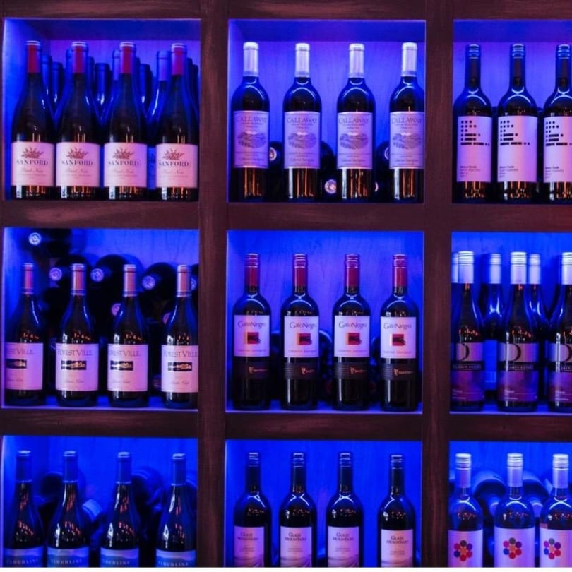 A wine display at Vino Cappuccino with multiple bottles of red wine illuminated by blue lights.