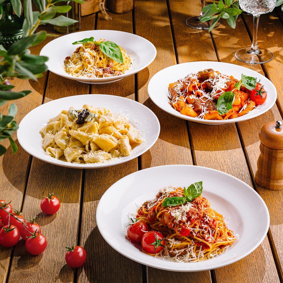 Four plates of various Italian pasta dishes, including spaghetti and penne, on a wooden table.