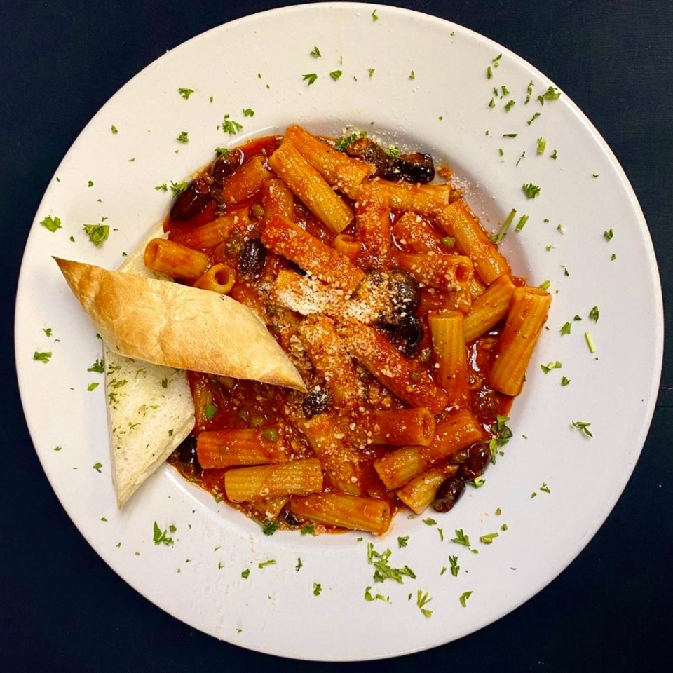 Rigatoni pasta in tomato sauce with olives, grated cheese, parsley, and toasted bread on a white plate.
