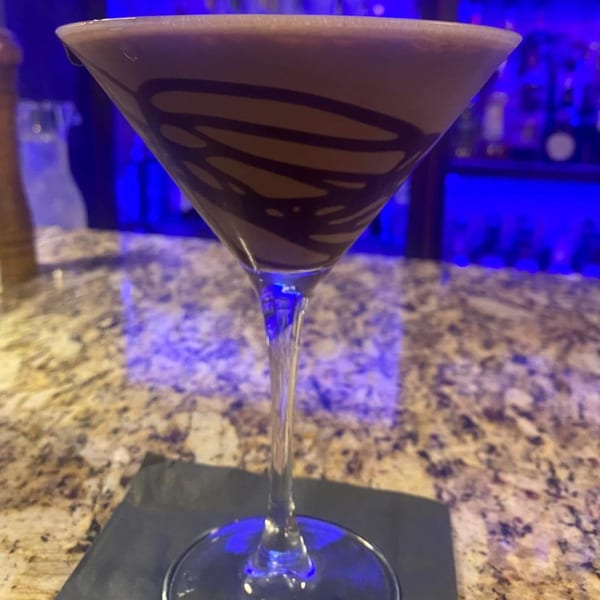 A chocolate martini with chocolate syrup swirls, on a bar with blue lighting.