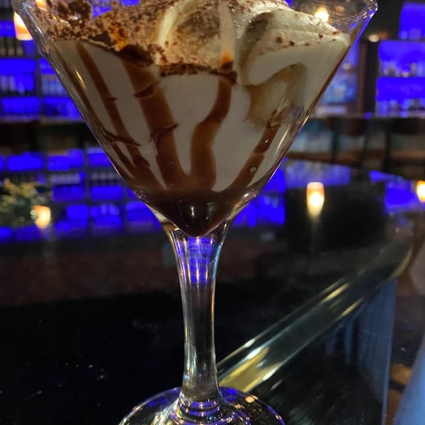 Martini glass filled with a creamy dessert, drizzled with chocolate, against a blue-lit background.