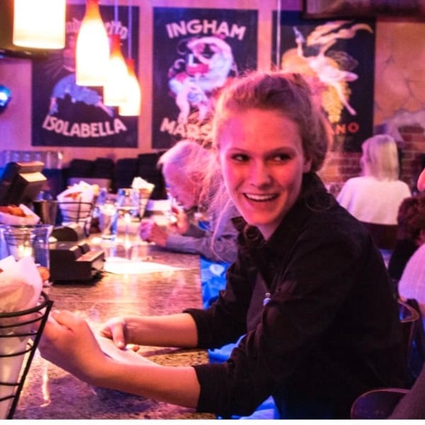 A smiling woman at a bar with vintage Italian posters and warm lighting in the background.