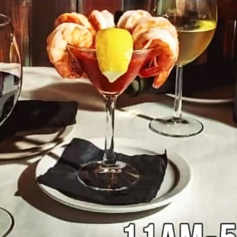 Shrimp cocktail with lemon garnish in a martini glass on a black napkin, beside a pale cocktail in a restaurant.