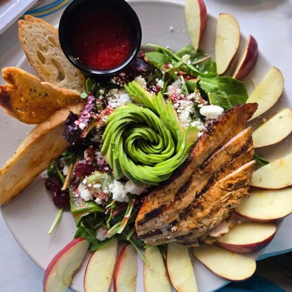 Grilled chicken salad with avocado, feta, cranberries, apples, and bread on a white plate.