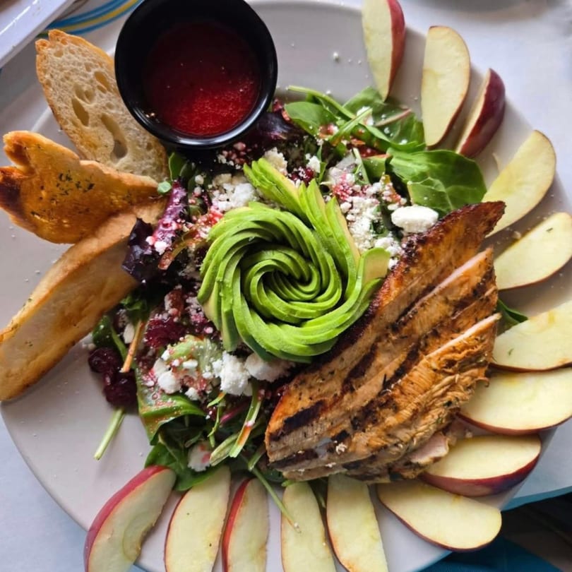 Grilled chicken salad with avocado, feta, cranberries, apples, and bread on a white plate.