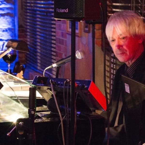 A musician with white hair plays a keyboard, illuminated by red and blue stage lights.